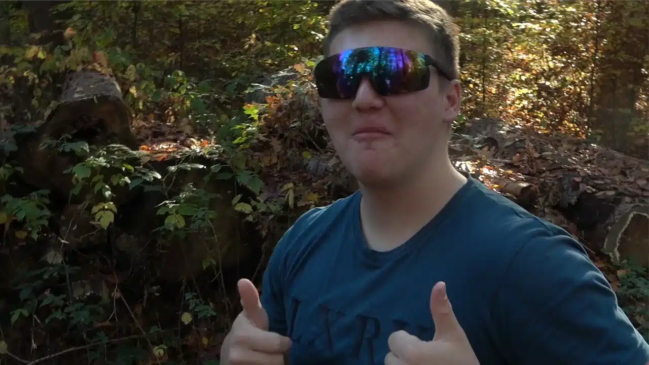 Ein junger Mann mit Sonnenbrille sieht witzig in die Kamera und zeigt beide Daumen nach oben