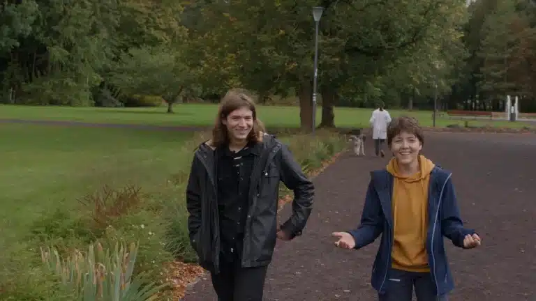 Zwei Personen laufen auf die Kamera zu und lachen sich an. Im Hintergrund verlässt eine Person in weißem Kittel das Bild.