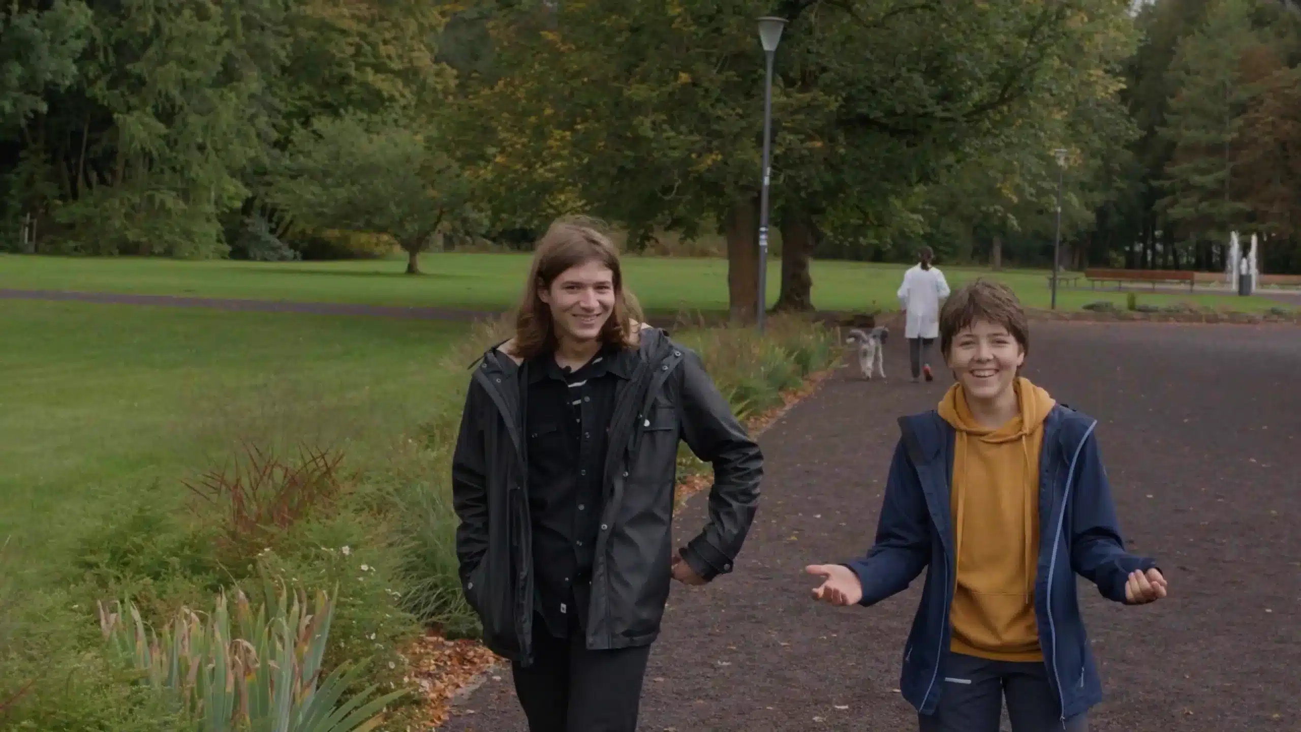 Zwei Personen laufen auf die Kamera zu und lachen sich an. Im Hintergrund verlässt eine Person in weißem Kittel das Bild.