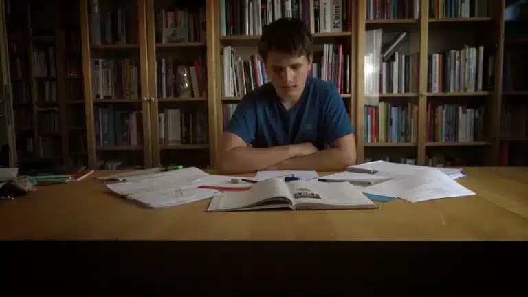 Ein Junge sitzt vor einem Bücherregal und hat viele Zettel um sich herum liegen.